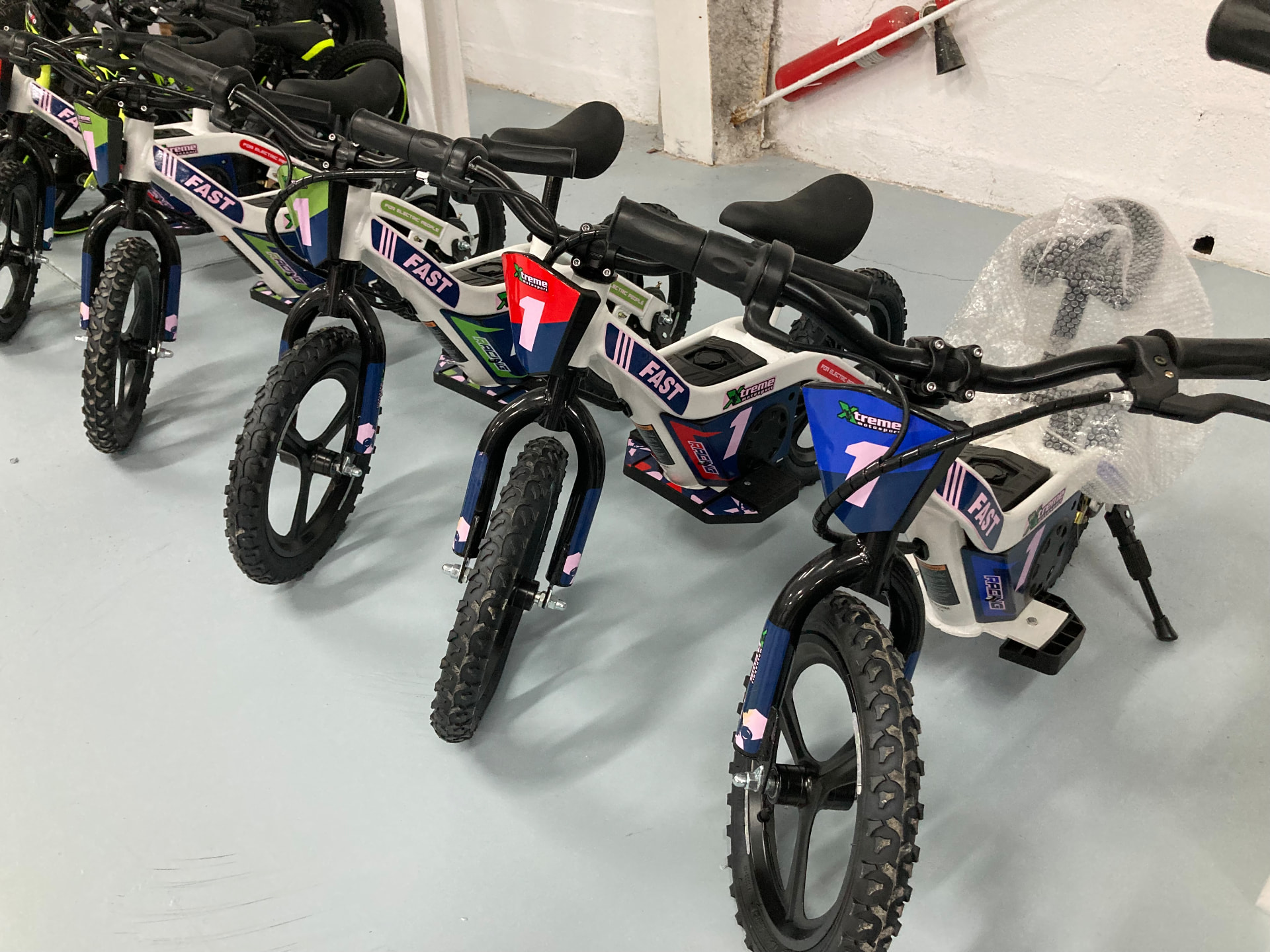 Bicicletas eléctricas infantiles Xtreme Motorsport con el dorsal 1 en blanco, azul y rosa alineadas en la nave