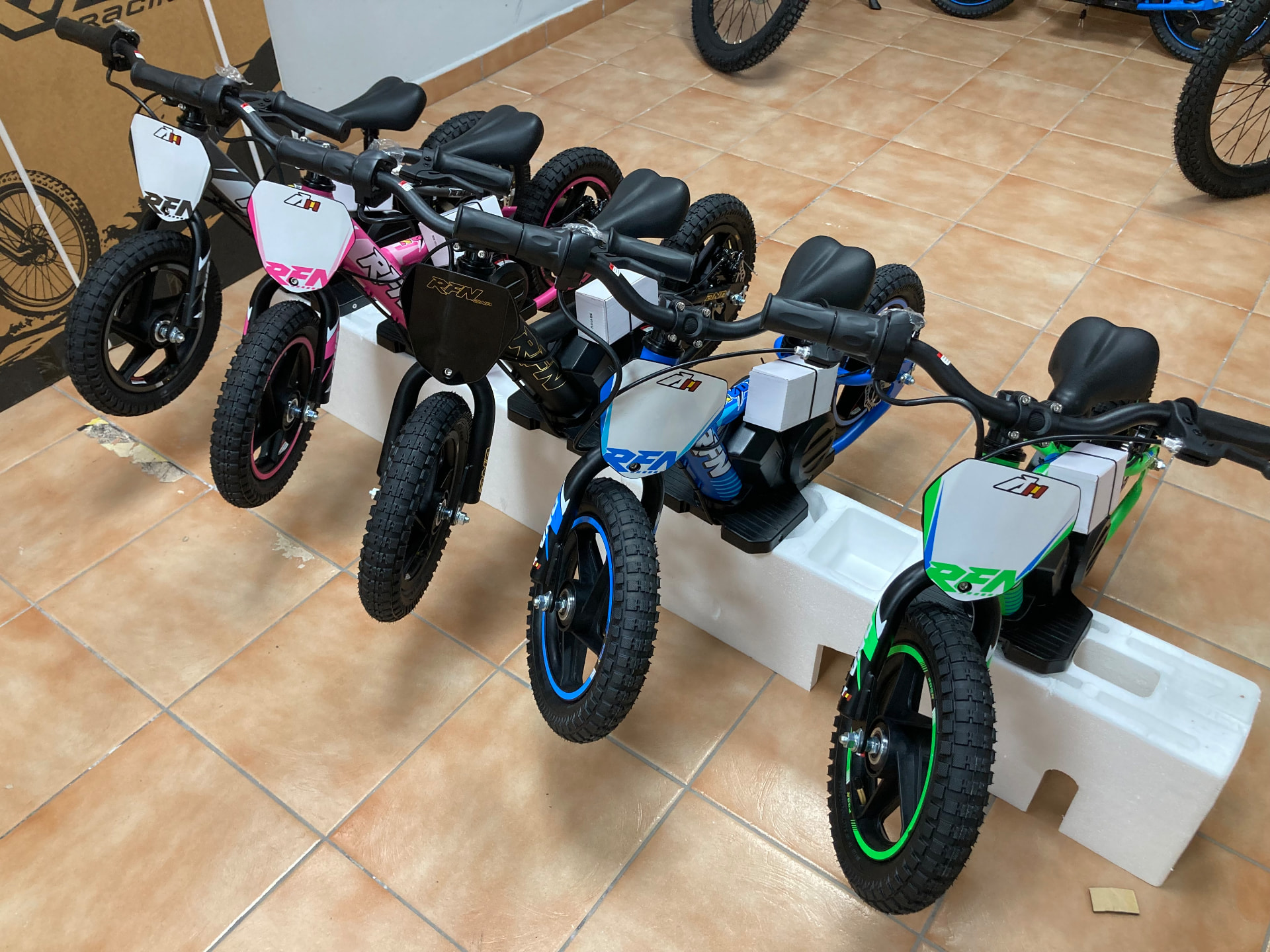 Fila de bicicletas eléctricas de equilibrio RFN infantiles en gris, rosa, negro, azul y verde lima sobre las bases de embalaje