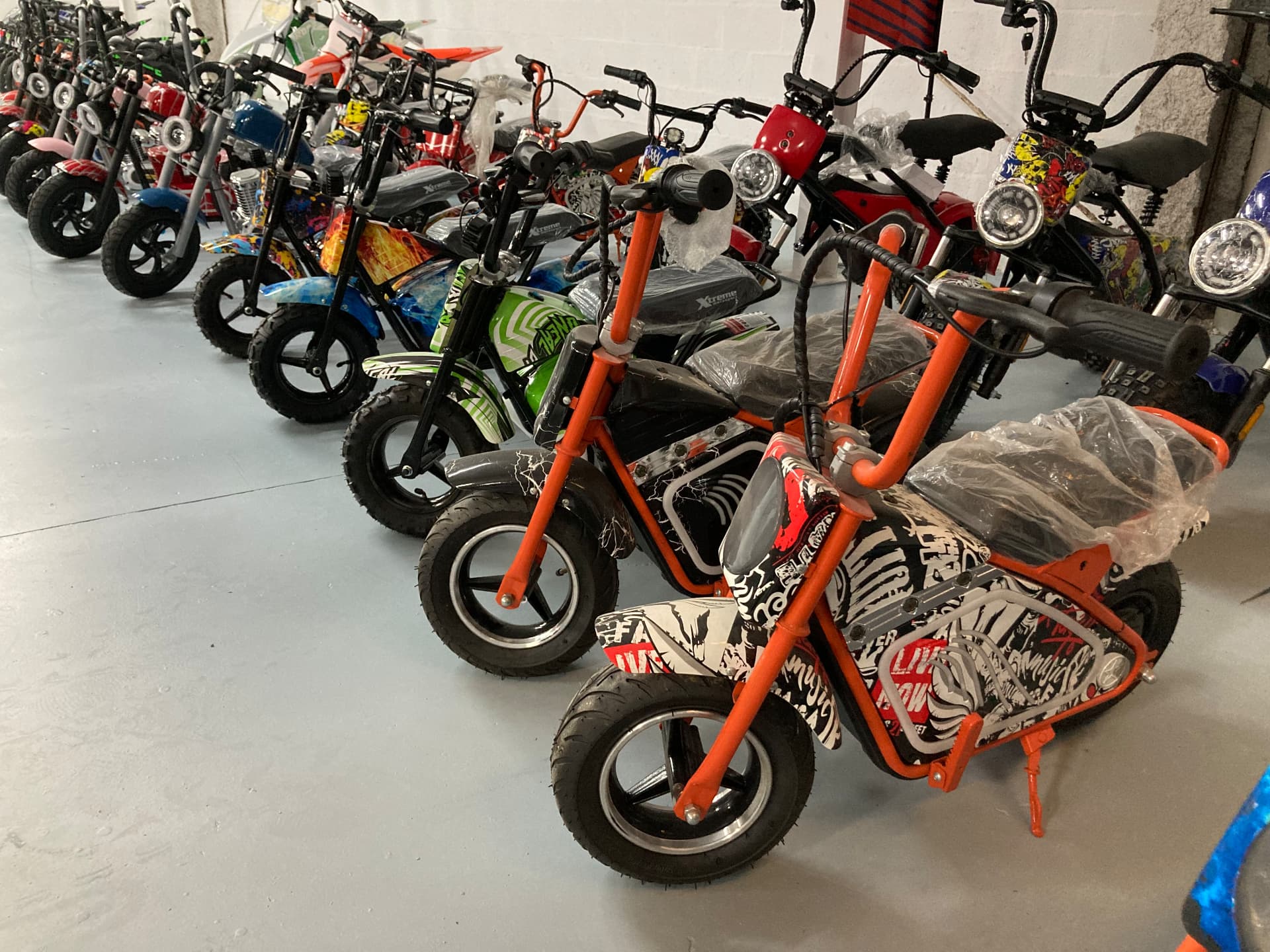 Fila de minimotos eléctricas infantiles Xtreme en naranja con gráficos de calavera, verde lima, azul con llamas, rojo, rosa y negro con frontal de cómic en el almacén